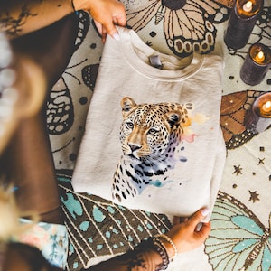 A person is holding a white t-shirt with a leopard print design on it, surrounded by candles and a colorful, patterned blanket.