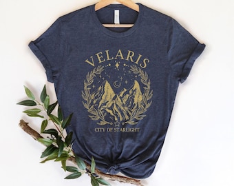 May include: A dark heather navy blue short-sleeve t-shirt featuring a gold graphic print of mountain peaks surrounded by a laurel wreath, stars, and a crescent moon. The text reads VELARIS and CITY OF STARLIGHT in a serif font.