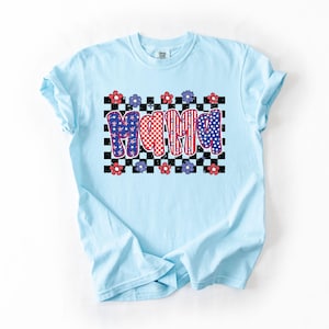 May include: Light blue t-shirt with the word "MAMA" in a patriotic design. The letters are decorated with red and white checkered patterns and blue stars. The shirt has a crew neck and short sleeves.