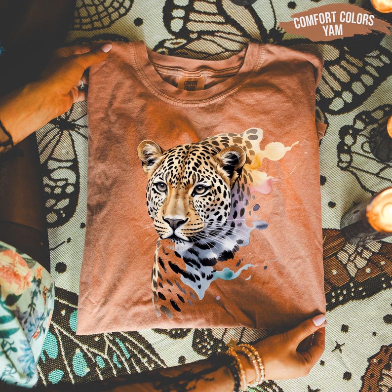 A person is holding a brown t-shirt with a leopard print design on it, which is placed on a patterned fabric.