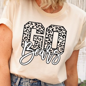 May include: A cream-colored short-sleeve t-shirt featuring a graphic design with the text "GO" in large block letters filled with a black and white leopard print pattern, and the word "Bears" written below in a white cursive script font.