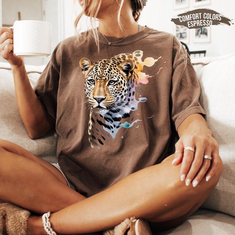A woman sitting on a couch, wearing a brown t-shirt with a leopard print design, holding a white mug and resting her hand on her knee.