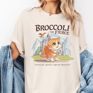 May include: A beige t-shirt with a graphic of a cartoon cat wearing a backpack and a dragon flying overhead. The text "Broccoli the Fierce" is above the cat and "Official Quest Squad Mascot" is below the cat.