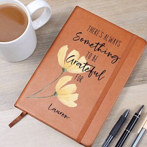 May include: A brown journal with the text "THERE'S ALWAYS SOMETHING TO BE GRATEFUL FOR" and a floral design. The journal has a ribbon bookmark and the name "Lauren" printed on it. A white mug of coffee, pens, and a phone are also visible.