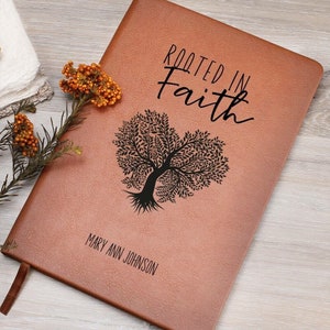 Może przedstawiać: Brązowy skórzany dziennik z tekstem "Rooted in Faith" i czarną sylwetką drzewa z korzeniami w kształcie serca. Imię "Mary Ann Johnson" jest spersonalizowane na dole dziennika.