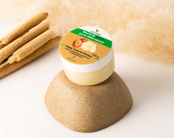 May include: A white jar with a green and yellow label that reads "Beurre de Kanité pur & 100% naturel" and "Visage, Corps et Cheveux". The jar is sitting on a brown bowl and contains a yellow shea butter product. The jar is 100 grams.