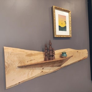 May include: A wooden shelf made from a single piece of wood with a natural edge. The shelf is mounted on a gray wall and has a small ceramic vase and wooden figurines on it. There is a framed picture above the shelf.