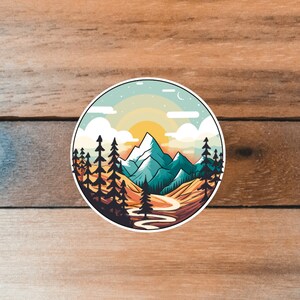 May include: A circular sticker with a mountain landscape illustration. The sticker features a blue sky with white clouds, a yellow sun, green mountains, a winding river, and a forest of dark green trees.