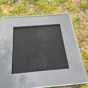 Op de afbeelding: Een grijze, rechthoekige vitrine met een zwart gaasscherm. De behuizing heeft een doorzichtige acryl behuizing en staat op gras. Het bovenste frame is effen grijs.