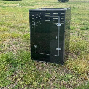 Op de afbeelding: Een zwarte acryl behuizing met een heldere acryl deur. De behuizing heeft ventilatiesleuven en is ontworpen om kleine dieren of reptielen te huisvesten. De structuur is rechthoekig en staat op een grasachtige ondergrond.