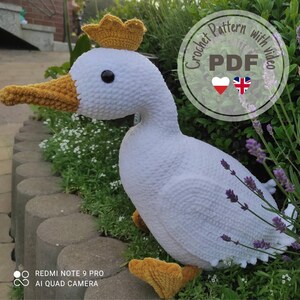 Könnte beinhalten: Eine große, weiße gehäkelte Gans mit goldener Krone, gelbem Schnabel und Füßen. Das Bild enthält den Text "Crochet Pattern with video PDF". Die Gans sitzt in einem Garten.