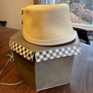 May include: A beige straw hat with a wide brim and a white ribbon around the crown. The hat is sitting in a brown box with a black and white checkered pattern.