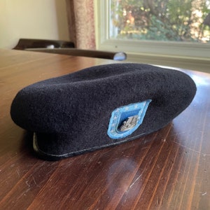 May include: A black beret with a blue patch featuring a white star and the number "11" in the center. The beret is made of wool and has a leather band around the bottom.