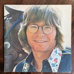 May include: A close-up portrait of a man with long, light brown hair wearing round glasses and a blue denim jacket with a white and floral patterned collar. The man is smiling and looking directly at the camera.