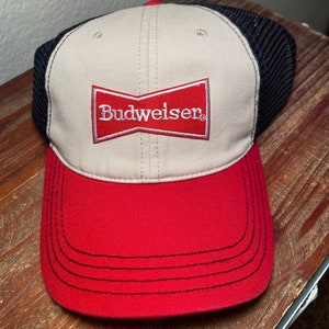 Puede incluir: Una gorra de béisbol beige y roja con una malla negra en la parte posterior. La gorra tiene un logotipo de Budweiser rojo bordado en la parte delantera.