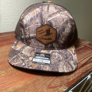 May include: Camouflage trucker hat with a brown leather patch that says "South Dakota" and features a pheasant. The hat is adjustable and has a mesh back.