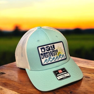 May include: A light blue trucker hat with a white mesh back and a patch that reads "Day Drinkin'" with a sun and wave design. The hat is made by Richardson and has a low-profile fit.