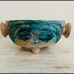 May include: A ceramic bowl with a face design. The bowl features a turquoise and tan color scheme, with a face sculpted into the front. The eyes are closed, and the ears are protruding from the sides. The bowl is resting on a white surface.