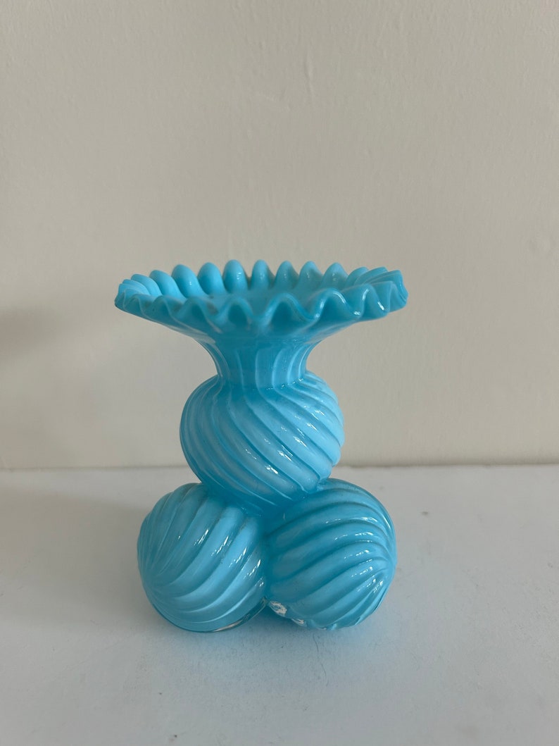 May include: A light blue glass vase with a ruffled top and a base made of three round, spiraled sections.