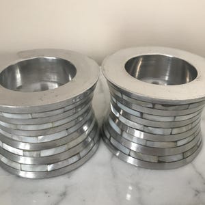 Puede incluir: Dos candelabros de plata con incrustaciones de nácar blanco. Los candelabros son cilíndricos y tienen una parte superior ligeramente acampanada.