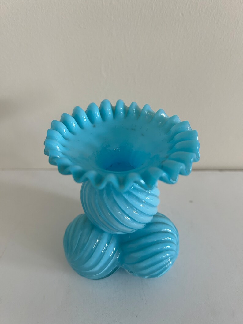 May include: A light blue glass vase with a ruffled top and a base made of three spiraled balls.