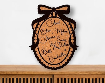 May include: An oval-shaped cork board featuring a black decorative frame with a bow at the top. The center of the board displays the names Ariel, Elsa, Mulan, Aurora, Snow White, Moana, Belle, and Cinderella in black script lettering with small star accents.