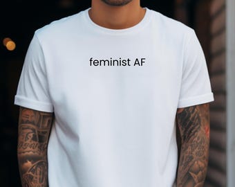 May include: A white short-sleeved crew neck t-shirt featuring the text "feminist AF" printed in black lettering across the center of the chest. The garment is made of a plain white fabric with a standard fit and simple hemmed sleeves.