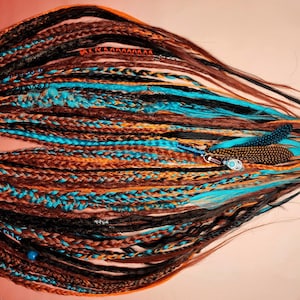 K&ouml;nnte beinhalten: Ein Set synthetischer Dreadlocks in verschiedenen Braunt&ouml;nen, Orange und Blau. Die Dreadlocks sind miteinander geflochten und haben Perlen und Federn angebracht.
