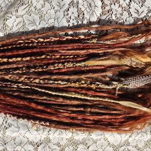 Puede incluir: Un conjunto de rastas sintéticas en tonos marrones, rojos y rubios. Las rastas están dispuestas en fila y tienen un aspecto natural y texturizado.