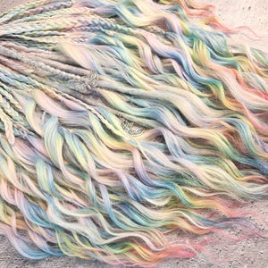 May include: Pastel-colored dreadlocks with a mix of braided and wavy sections. The color palette includes shades of pink, blue, yellow, and green. Silver butterfly and crescent moon charms are attached to the hair. The hair is styled on a neutral surface.