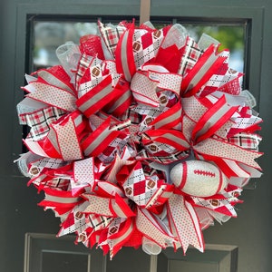 May include: A red, white, and gray football wreath with a silver football in the center. The wreath is made of ribbon and mesh and features a plaid pattern, polka dots, and football designs.