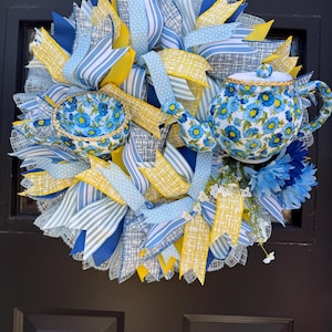 May include: A blue and yellow wreath with a teapot and teacup, decorated with blue and white floral patterned fabric. The wreath is made with burlap, ribbon, and mesh.
