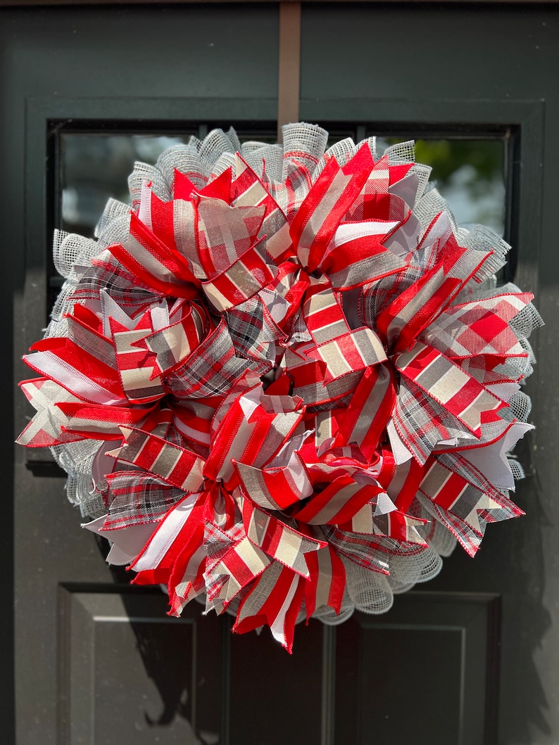 May include: A red, white, and gray plaid and striped wreath with a white mesh base. The wreath is decorated with multiple bows and is perfect for a front door.
