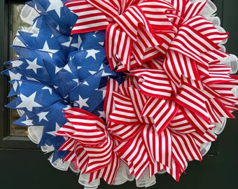 May include: A patriotic wreath featuring a design split into two halves. One side showcases blue ribbon with white stars, the other red and white striped ribbon. The wreath is trimmed with white mesh.