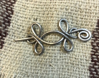 May include: A silver metal Celtic knot pin with a decorative swirl at the end. The pin is on a brown and beige woven fabric.
