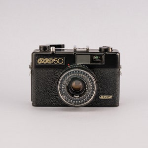 Op de afbeelding: Een zwarte 35mm filmcamera met een gouden inscriptie die "Smena 50" en "Автомат" aangeeft.