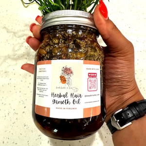 May include: A glass jar filled with herbal oil for hair growth, held by a person with red nail polish. The jar has a silver lid and a label that reads "Herbal Hair Growth Oil, Made in Virginia." Green plants are placed on top of the jar.