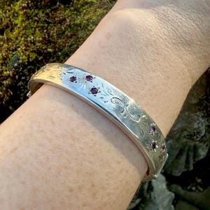 May include: A silver hinged bangle bracelet, featuring engraved floral patterns and small, round, dark red gemstones. The bracelet is worn on a wrist, highlighting its design. The bracelet is a piece of jewellery.