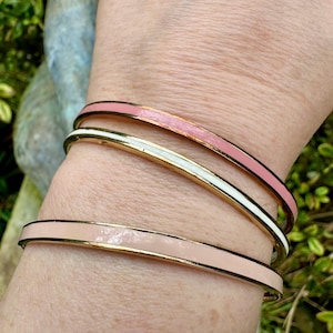 Può includere: Tre braccialetti dorati con smalto rosa e bianco. I braccialetti sono impilati su un polso, mostrando i diversi colori e design. I braccialetti hanno una finitura lucida e un design semplice ed elegante.