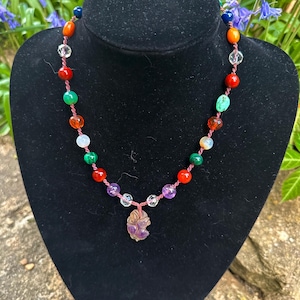 May include: A multi-coloured beaded necklace with a carved amethyst pendant. The necklace features beads in shades of red, orange, yellow, green, blue, and purple.