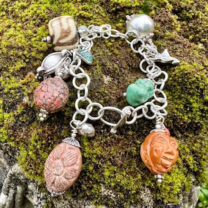 May include: A silver charm bracelet with a variety of charms, including a carved wooden bead, a turquoise stone, and a carved stone face. The bracelet is on a mossy green background.