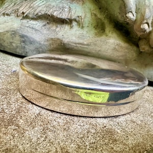 May include: A polished, oval-shaped silver box with a smooth, reflective surface. The lid features a small, engraved hallmark near the center edge, indicating its metallic composition. The container has a simple, rounded design with a seamless closure.