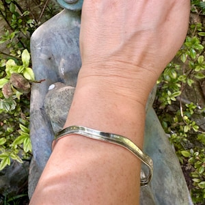 May include: A silver-toned metal cuff bracelet with a smooth, undulating wave-like design. The bracelet features a polished, reflective surface that follows a gentle, non-linear path around the wrist, showcasing a simple and clean aesthetic.