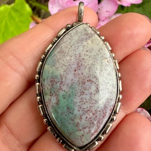 May include: A gemstone pendant with a silver-coloured metal frame. The stone has a light green and red speckled pattern. The pendant is held in a hand, with pink flowers in the background.
