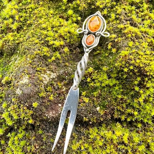May include: A silver-colored, decorative fork with a twisted handle and two amber-colored stones. The fork is set against a backdrop of green moss, creating a contrast between the metal and the natural environment.
