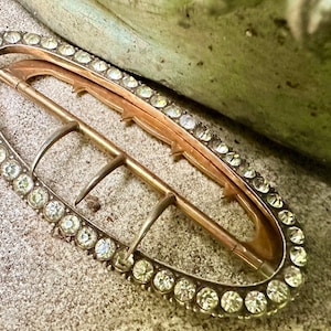 May include: An oval-shaped metal buckle with a decorative row of clear rhinestones around the edge. The buckle has a gold finish and a pronged closure.
