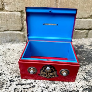 May include: A red metal box with a blue interior and a hinged lid. The box features two silver-colored knobs and a decorative emblem with the word "Royal". The box is open, revealing the blue interior.