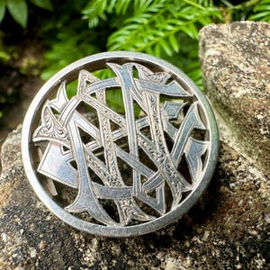 May include: A silver circular brooch with an elaborate, interwoven design. The brooch displays a series of stylized letters or symbols, forming a complex pattern. The brooch is set against a natural, textured stone background.