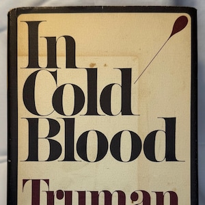 May include: Vintage hardcover book titled "In Cold Blood" by Truman Capote. The book cover features bold black lettering for the title and maroon lettering for the author's name. A maroon teardrop graphic is above the title.