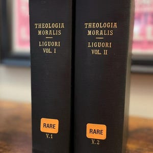 May include: Two hardcover books with black covers. The text "THEOLOGIA MORALIS LIGUORI VOL. I" and "THEOLOGIA MORALIS LIGUORI VOL. II" are printed in gold. Each book has an orange sticker that reads "RARE" and "V.1" or "V.2".